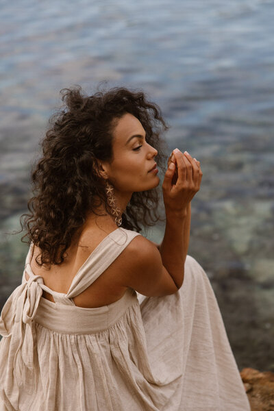 A woman wearing a dress with curly hair is sitting with her back to the camera in front of the sea.