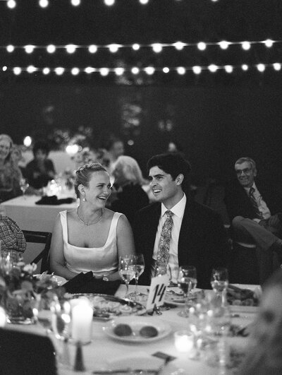 A bride and groom sitting at their reception table looking at eachother and reacting to some speeches. There are wine glasses and food on the table, and more wedding guests behind them. There are market lights above them, and the photo is a black and white film image