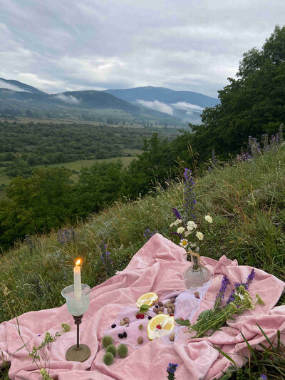 Idyllic picnic setup with fruits, candles, and satin blanket in front of a mountain landscape.
