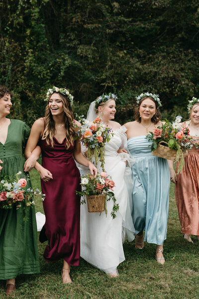 Bride walking outdoors with her bridesmaids, all wearing colorful mismatched dresses and floral crowns, linked arm-in-arm while holding lush garden-style bouquets featuring roses, dahlias, delphinium, ranunculus, and greenery. The group is smiling as they walk across a grassy field with tall trees in the background, showcasing vibrant floral design and whimsical wedding party styling.