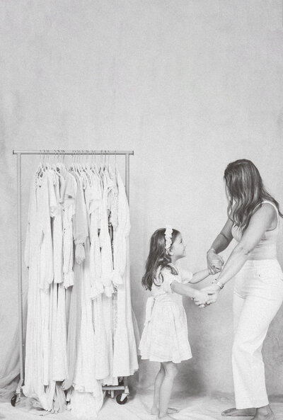 Smiling daughter holding hands with her mother while standing in front of a curated wardrobe rack during a studio photo session.