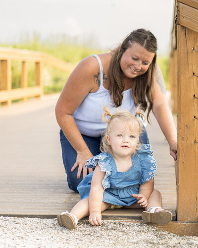 Mom and daughter sitting on a bridge at a wildlife sanctuary.  Mom is a brunette, wearing a white tank top, blue jeans, smiling down at her daughter.  Daughter has blond hair wearing a blue dress and sandals, smiling at the camera