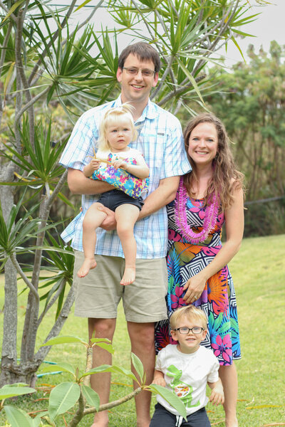 A family with two young toddlers standing together in front of some tropical foliage.