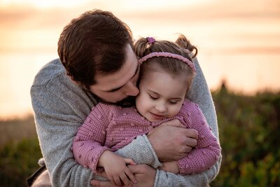 Dad cuddling his young daughter on the beach dunes with the sun setting behind