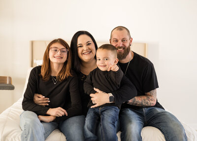 Family of five sitting on a bed looking at the camera smiling.  All are wearing black shirts and blue jeans.  Daughter is wearing glasses