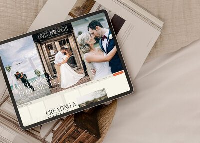 A tablet displaying a wedding photography website, crafted by an expert Showit website designer for photographers, lies on top of an open magazine on a beige fabric surface.