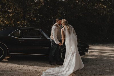 Bride and groom standing outside kissing in front of old car