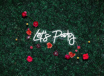 A neon sign that says lets party on a greenery wall with colorful flowers around it