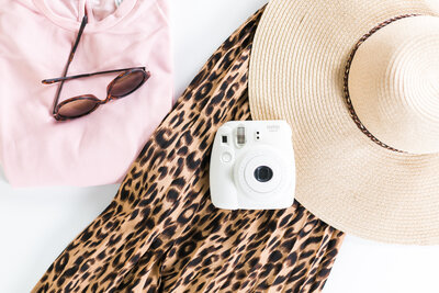 aerial view of a pink shirt, sunglasses, a straw hat, a leopard print skirt, and a white instax camera, getting packed for travel