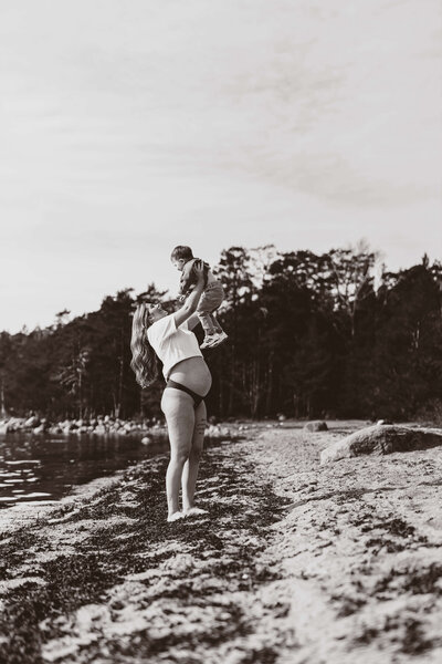 En svartvit bild på en gravid kvinna hållandes i hennes son uppe i luften ståendes på en strandkant under en gravidfotografering i Stockholm