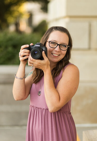Photographer with long dark brown hair, black glasses, smiling at the camera while holding a black camera, wearing a purple dress.