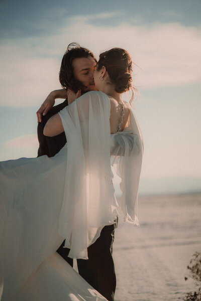 A groom holds a bride in his arms with the sunlight filtering through her dress on the Utah, Salt Flats.