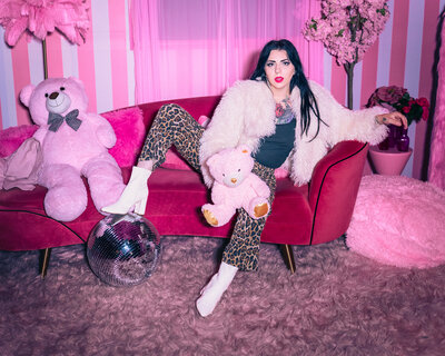 Editorial branding portrait by Moonlit Photo inside a vibrant pink studio near Salt Lake City, featuring a woman lounging on a velvet sofa with plush teddy bears, a disco ball, and retro-inspired styling. Bold, creative Utah branding photography with colorful, cinematic aesthetic.