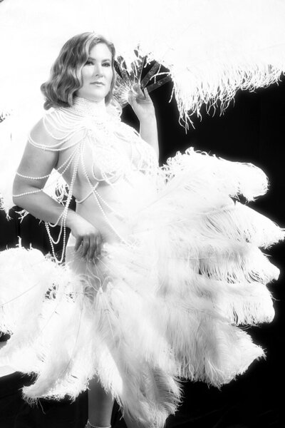 A woman is draped in pearls with white feather fans and photographed like a burlesque performer like Christina Aguilera.