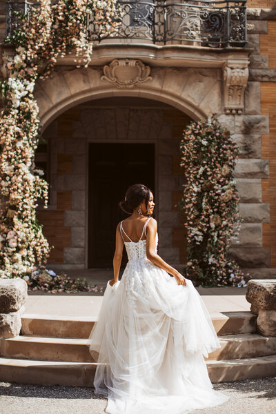 Bride in a flowing lace gown walking up the steps of a historic stone wedding venue decorated with flowers in Maine.