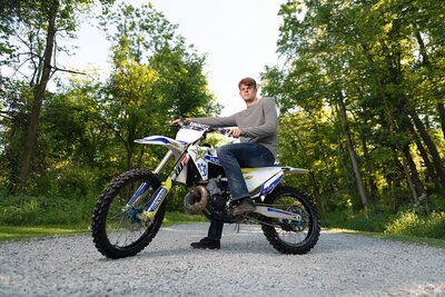 High school senior boy sitting on motorcycle on gravel path lined with trees