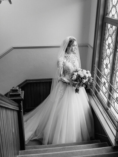 Bride holding bouquet and looking out stained glass window before wedding at Holy Angels in Sidney, Ohio