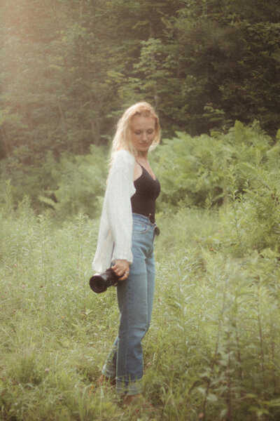 vermont photographer holding Canon camera in green grassy field, she's wearing blue jeans, a black tank top, and a white long sleeve shirt and has bright blonde hair