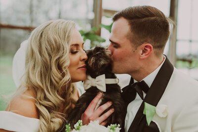 In a tender scene captured by a Columbus wedding photographer, a bride and groom lovingly kiss their small black dog, who is adorned with a charming white bow tie. The bride's long, wavy blonde hair and elegant makeup complement her radiant smile as she gently closes her eyes, while the groom, in a sharp black tuxedo, shows affection to their beloved pet. This heartwarming moment reflects the joy and love shared on their special day, surrounded by lush greenery in a bright conservatory setting
