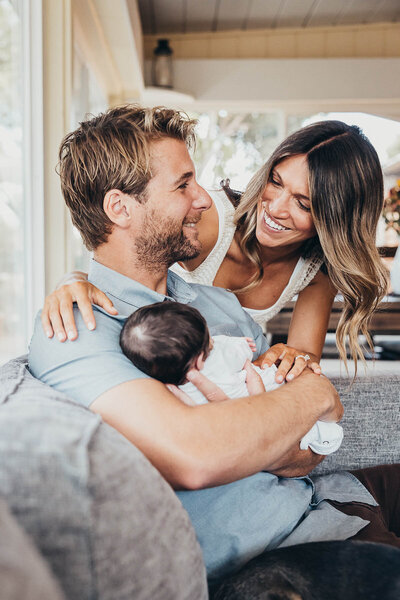 A father holds his newborn baby daughter while mom hugs them both and they look happily at each other in their home in San Diego, CA