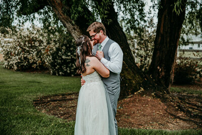 Bride and groom share an intimate moment under a tree during their outdoor wedding, standing close with their foreheads touching