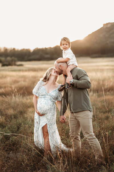 family walking through a field with son on dad's shoulders and mom kissing dad