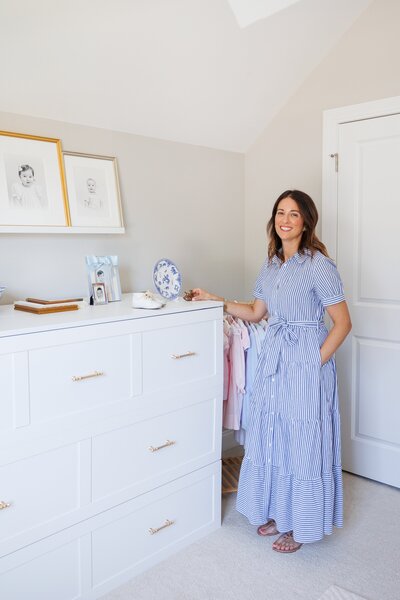Woman in a striped dress standing in a bright room beside a dresser and children’s clothes.