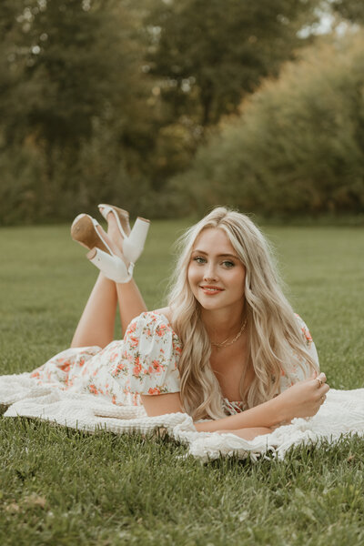 Senior portrait session by Bailee Freeman Photography for a blonde model with long hair. The model is wearing a pink and cream floral dress against a field of green grass and green trees in the distance. 