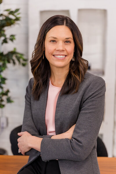 woman with long brown hear leaning against a desk arms crossed wearing corporate outfit light pink top and gray jacket during headshot photoshoot with Laure Photography