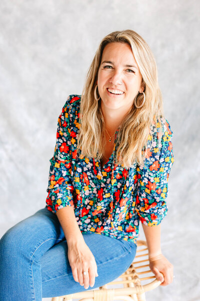 Blond woman in a floral shirt and blue jeans casually sitting on a wooden stool leaning to one side and smiling against a gray backdrop