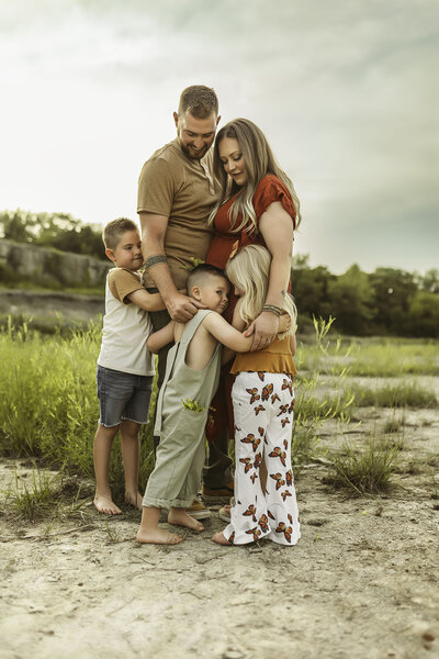 Mom in red dress hugs husband in brown shirt while both look down at their three children in a candid moment outdoors
