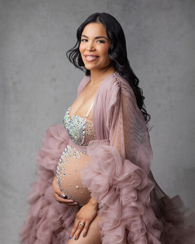 Portrait of pregnant dark - haired woman standing and posed at a 45 degree angle, looking and smiling with her teeth.  She is posed against a textured gray backdrop and is wearing a nude bodysuit with rhinestones over her chest and down the middle of her belly.  She is also wearing a mauve colored tulle robe with full ruffles on her sleeves and midway to bottom of her robe