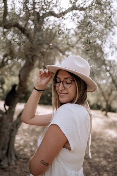 Oberkörper Foto von Frau (Lisa Winkelbauer) Sie trägt ein weißes Oberteil und einen beigen Hut. Sie lächelt, ihre Augen sind geschlossen und der Kopf leicht nach unten geneigt.
