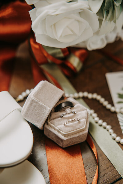 Flat lay of rings in a white velvet box on ribbon, with the bride's shoe and bouquet in view.