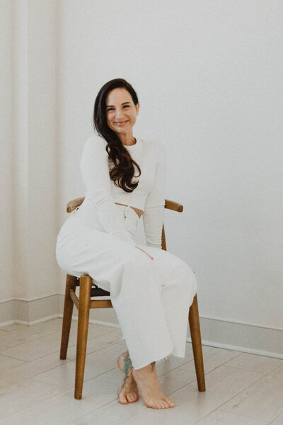 Holistic Esthetician and Online Skincare Specialist, Jen, sitting in a wood chair wearing a white top and pants