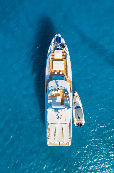 Aerial view of a large white yacht anchored in clear blue water, with a small boat tied alongside.