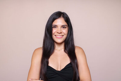 Woman with long black hair posing in front of a soft blush pink background.