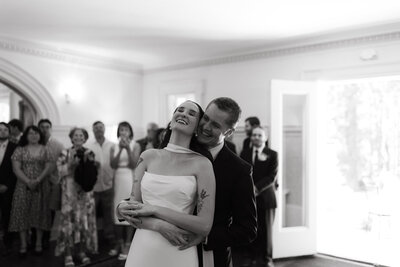 A documentary style photos of the first dance between a newly wed couple. The groom hugs the bride from behind and she falls into his embrace. 