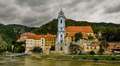 Riverside view of Dürnstein, Austria, featuring the town’s blue Baroque church tower, colorful buildings, and surrounding green hills along the Danube River.