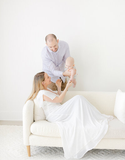 young woman wearing simple white dress sits on the couch looking up at baby boy held by husband