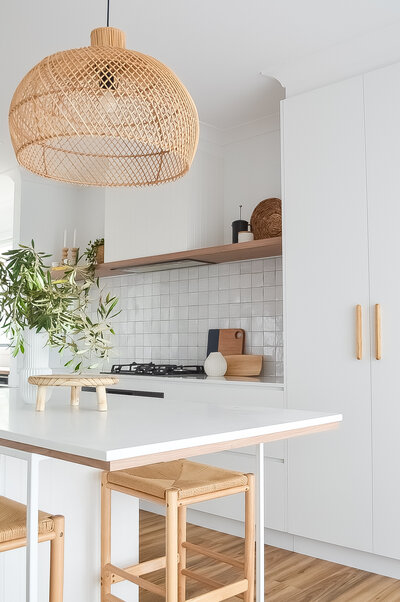 Minimalist kitchen featuring a rattan pendant light, white cabinets, tiled backsplash, and wooden stools—an exquisite touch by a Newcastle interior designer.