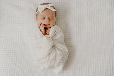 baby girl wearing white bow wrapped in white blanket on white bed in Annapolis Maryland