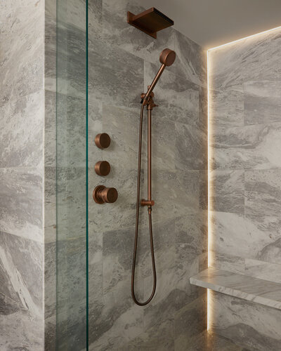 A walk-in shower featuring marble walls, flooring, and shelving, bronze shower fixtures, and warm backlighting