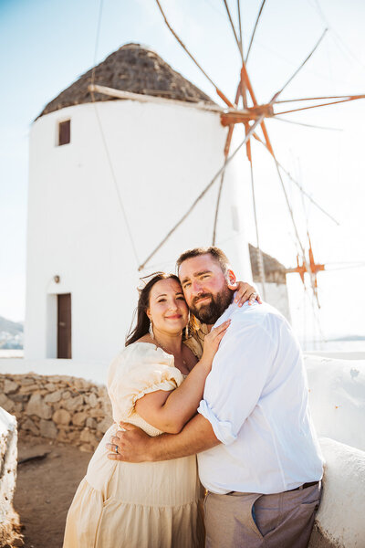 A couple smiling at the camera in mykonos greece with a windmill in the background.