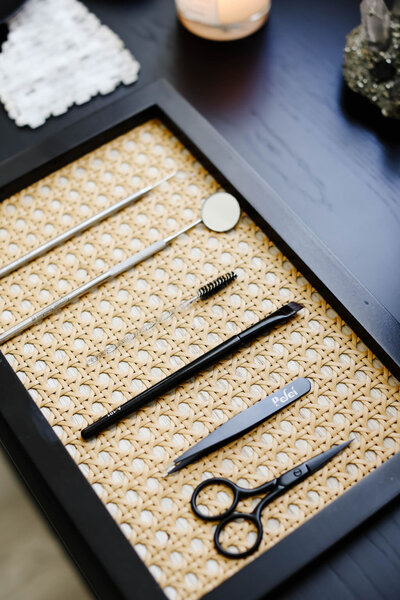 A neatly arranged set of professional brow and lash tools, including tweezers, scissors, spoolie, and other precision instruments on a woven mat, showcasing the high-quality tools used for brow lamination and lash lift services at Skin Ritual Co