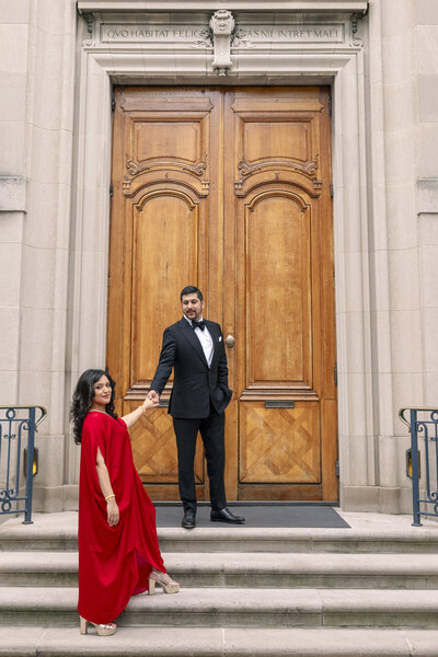 A couple stands on the steps of a building with an ornate wooden door. The woman in a flowing red dress holds hands with the man in a black tuxedo as they look at each other. Latin text is engraved above the door.
