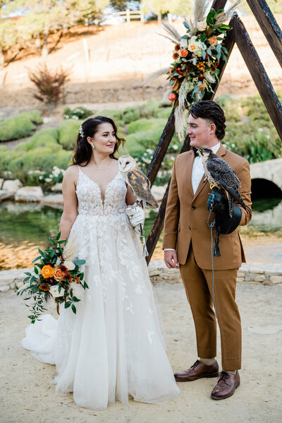 A bride and Groom at Terra Mia Vineyards stand in front of their bohemian wedding arch in November, and fall wedding colors, holding a hawk and a barn owl that were their ringbearer in a whimsical and playful wedding day ceremony.