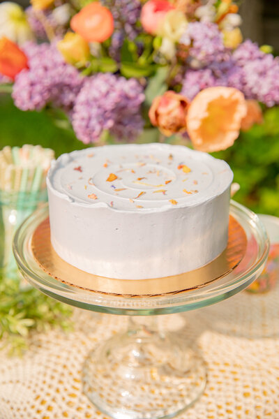 lavendar sweet heather anne cake on class stand with flowers in the background during styled shoot