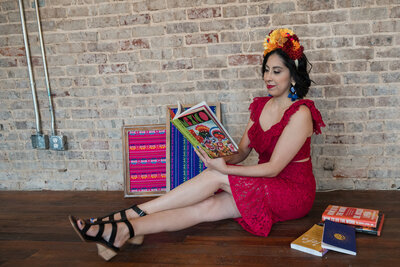 The artistic world as the founder of our art workshops sits on the floor in a captivating red lace dress, holding a book with a vibrant cover. Behind her, witness the beauty of handmade paintings crafted in our workshops.