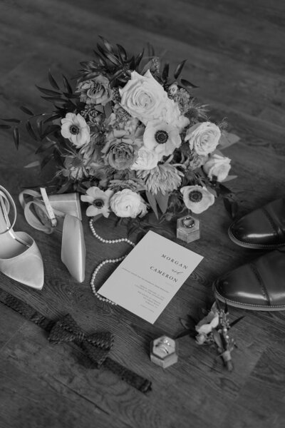Wedding flat lay items arranged on a wooden floor including a bridal bouquet, heels, men's dress shoes, a wedding invitation, a pearl necklace, rings in boxes, a bowtie, and a boutonniere.
